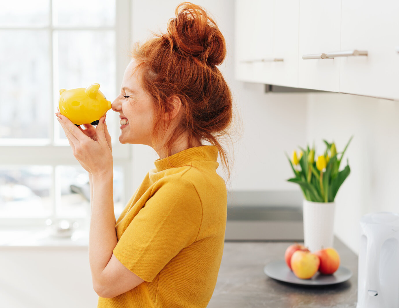 Smiling red-haired woman playing with yellow piggy bank in her hands, viewed from the side in bright room interior. Money savings concept