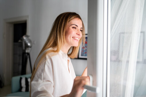 Beautiful smiling young woman opening window at the morning, fresh air, 