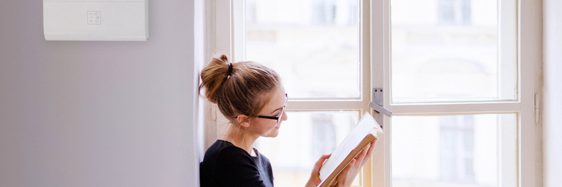 A young happy college female student with a book sitting on window sill at home, studying.